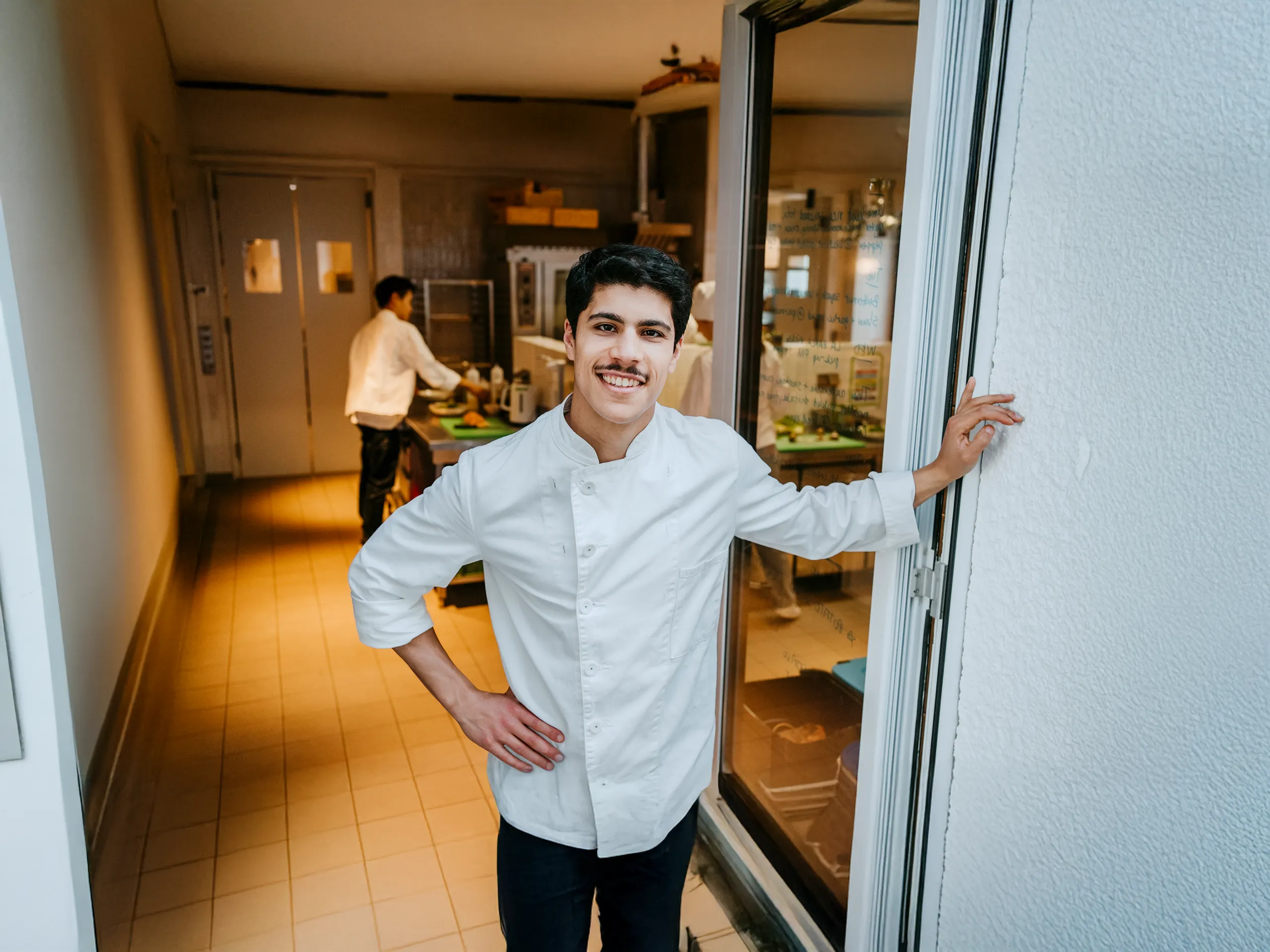 Young chef standing and smiling in front of a restaurant kitchen.