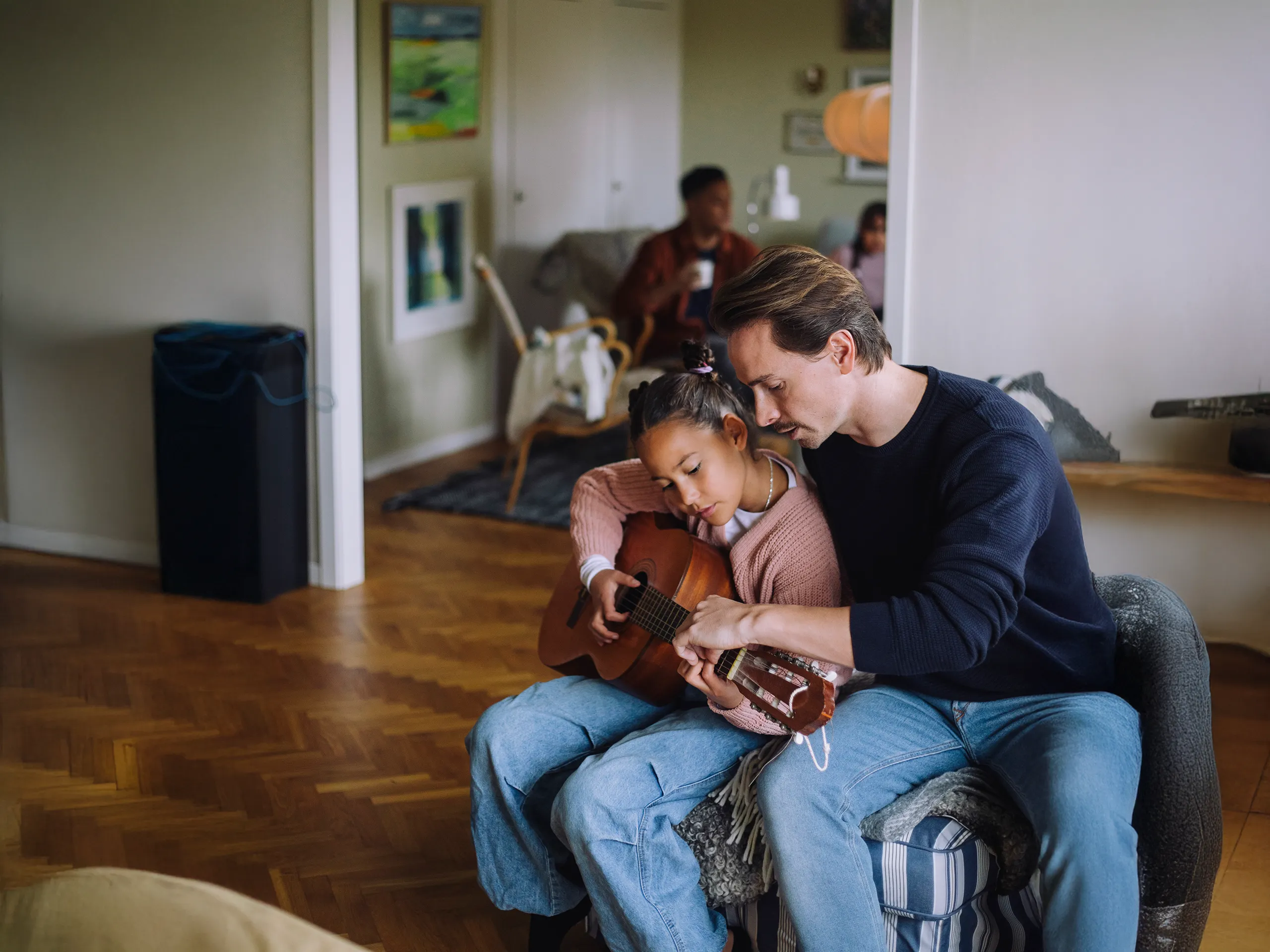 Father teaching his daughter how to play the guitar in a living room.
