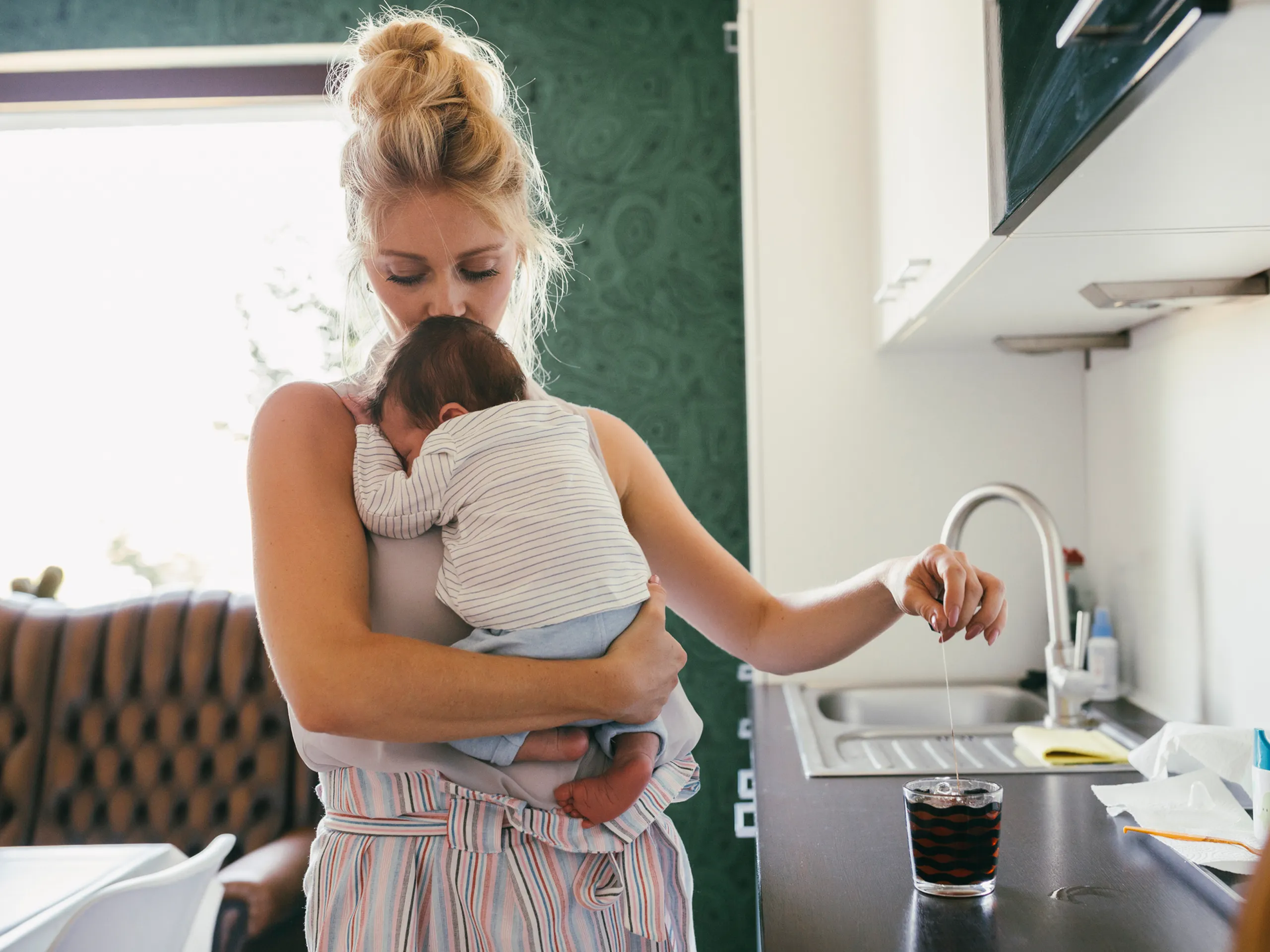 Mother holds her baby while preparing tea in the kitchen.