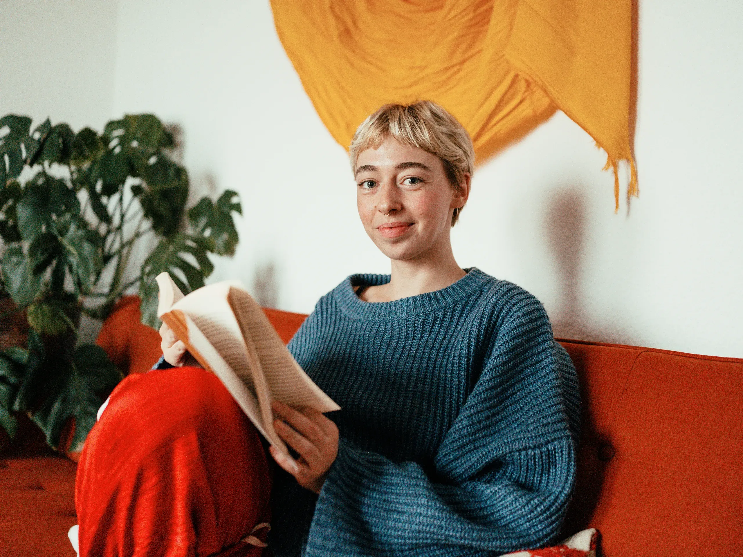 Smiling woman with short blonde hair reading a book on a red sofa.